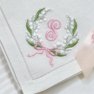 May include: White linen napkin with a delicate embroidered design. A wreath of white flowers and green leaves surrounds a pink letter "S". A pink bow is at the bottom of the wreath. The napkin has a stitched border.
