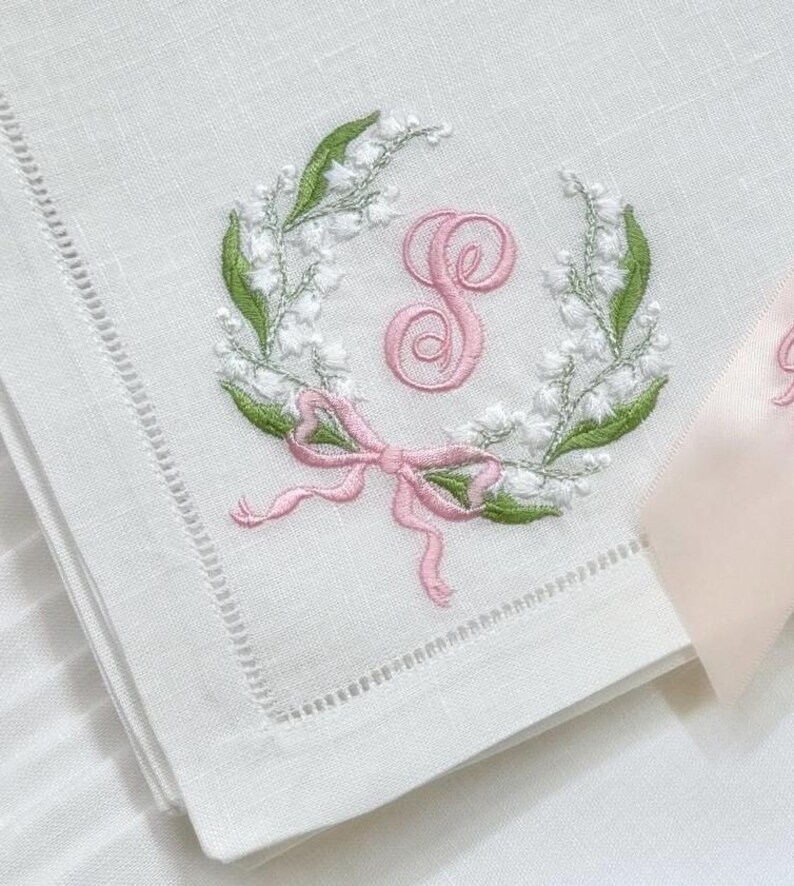 May include: White linen napkin with a delicate embroidered design. A wreath of white flowers and green leaves surrounds a pink letter "S". A pink bow is at the bottom of the wreath. The napkin has a stitched border.