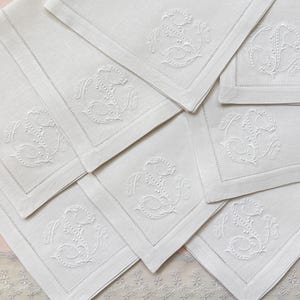 May include: A collection of white linen napkins, each embroidered with a monogram. The napkins are square with a detailed border stitch. The embroidered monogram features the letters "H" and "S" in a decorative script.
