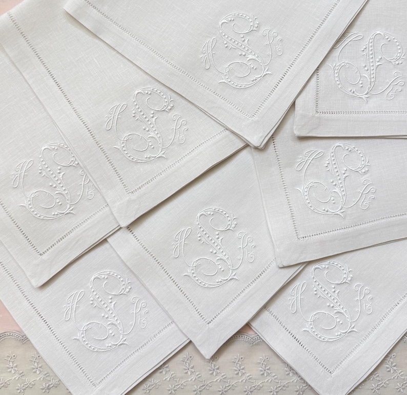 May include: A collection of white linen napkins, each embroidered with a monogram. The napkins are square with a detailed border stitch. The embroidered monogram features the letters "H" and "S" in a decorative script.