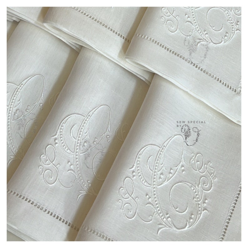May include: A stack of ivory-colored linen napkins. Each napkin features an embroidered monogram with ornate, swirling details. The edges are finished with a delicate, stitched border. The napkins are neatly folded, showcasing the elegant design.
