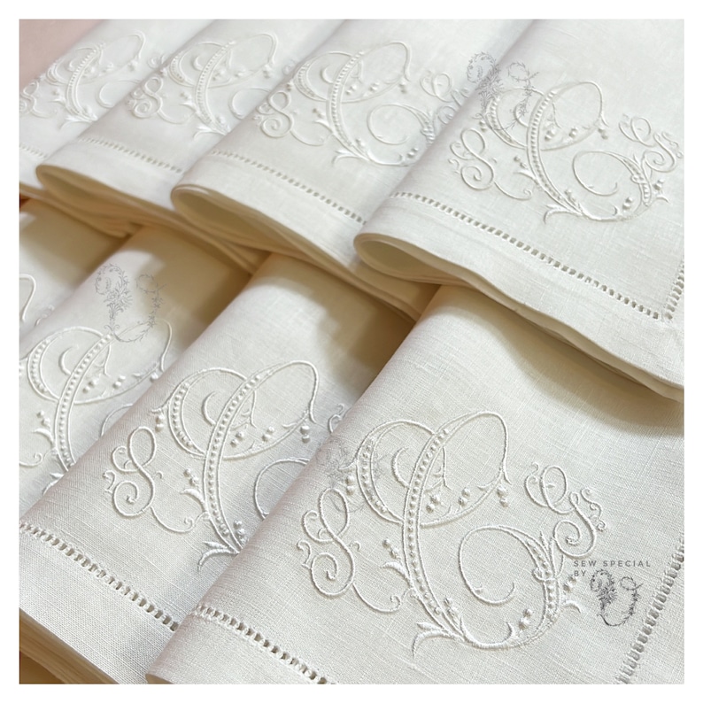 May include: A stack of off-white linen napkins, each embroidered with a large, ornate "CS" monogram. The napkins have a delicate, stitched border. The image is well-lit, highlighting the texture and detail of the embroidery.