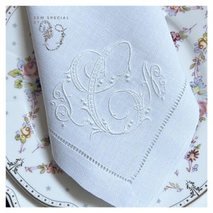 May include: White linen napkin with embroidered initials. The napkin features a decorative monogram in white thread, with a delicate floral design. The napkin is placed on a floral patterned plate. The text "SEW SPECIAL BY" is in the upper left corner.