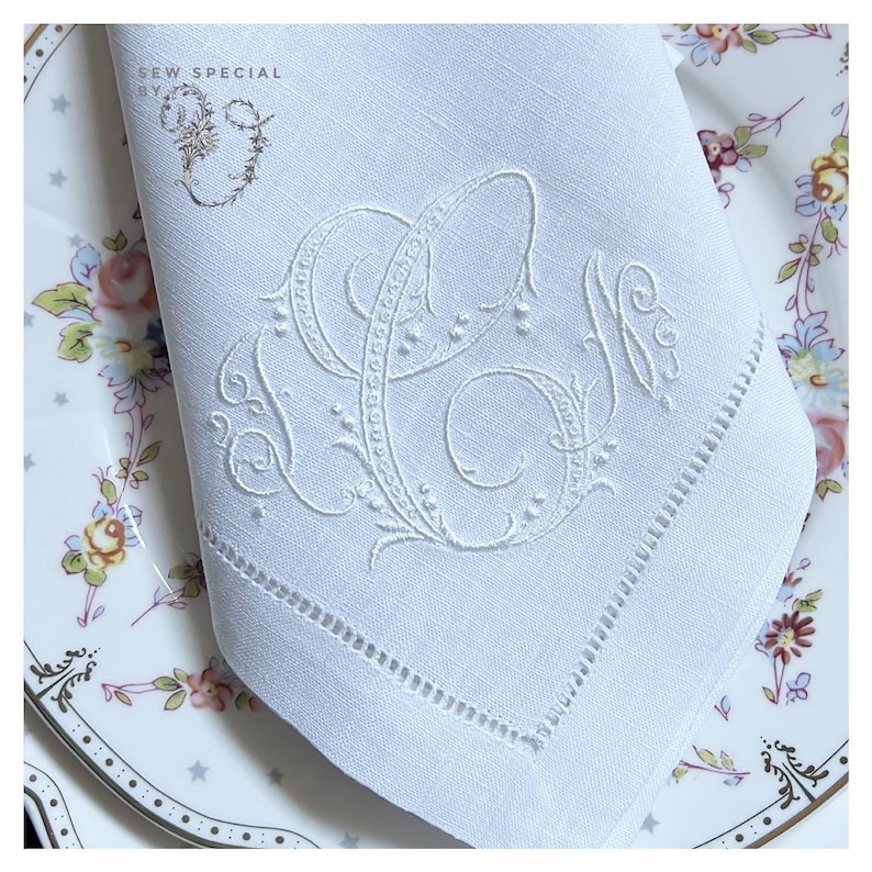 May include: White linen napkin with embroidered initials. The napkin features a decorative monogram in white thread, with a delicate floral design. The napkin is placed on a floral patterned plate. The text "SEW SPECIAL BY" is in the upper left corner.