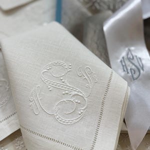 May include: White linen napkin with an embroidered letter "S" in a decorative script. The napkin has a stitched border and is folded. A white ribbon with embroidered initials is in the background.