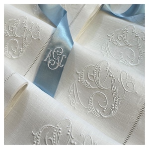 May include: White linen napkins with embroidered initials and decorative flourishes. A light blue satin ribbon with embroidered initials is wrapped around the napkins. The napkins have a delicate, elegant appearance.