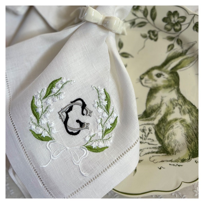 May include: White linen napkin with embroidered lily of the valley and a monogram. A white ceramic bow napkin ring holds the napkin. A decorative plate with a green rabbit illustration is in the background.