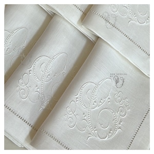 May include: Close-up of several folded white linen napkins. Each napkin features an embroidered monogram with the letters "S" and "C" in an elegant, ornate script. The napkins have a delicate, hemstitched border. The overall aesthetic is classic and refined.