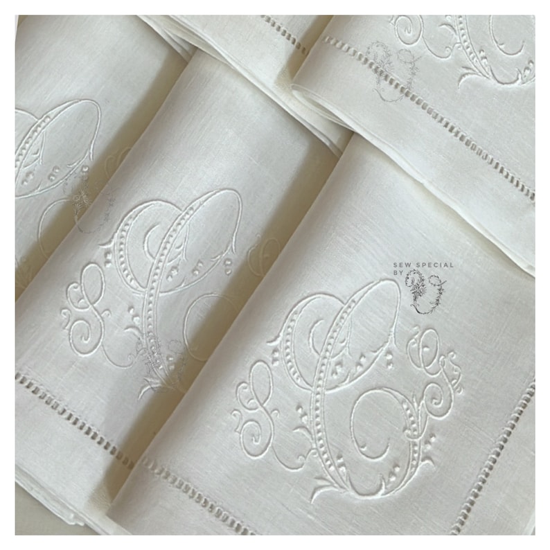 May include: Close-up of several folded white linen napkins. Each napkin features an embroidered monogram with the letters "S" and "C" in an elegant, ornate script. The napkins have a delicate, hemstitched border. The overall aesthetic is classic and refined.