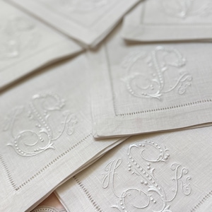 May include: Close-up of several white linen napkins with embroidered initials. The napkins have a delicate, detailed design with a decorative border. The initials are in a classic, elegant script.
