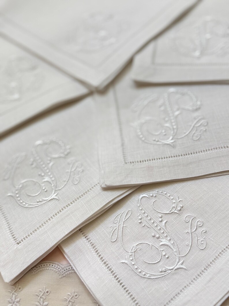 May include: Close-up of several white linen napkins with embroidered initials. The napkins have a delicate, detailed design with a decorative border. The initials are in a classic, elegant script.