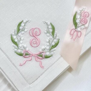 May include: White linen handkerchief with embroidered floral wreath, pink bow, and initial "S". A matching ribbon with floral and initial embroidery is also present. The embroidery features green leaves and white flowers.