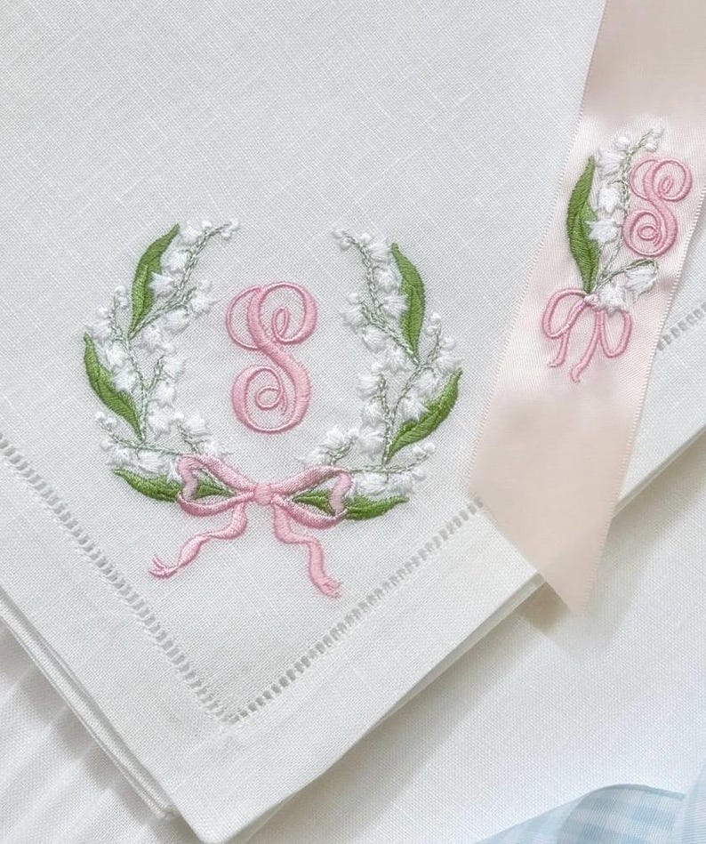 May include: White linen handkerchief with embroidered floral wreath, pink bow, and initial "S". A matching ribbon with floral and initial embroidery is also present. The embroidery features green leaves and white flowers.