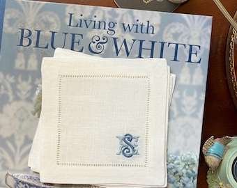 May include: A stack of white linen napkins with a blue embroidered initial "S" rests on a book titled "Living with BLUE & WHITE." The napkins have a decorative stitched border. A coaster and a small bell are also visible.