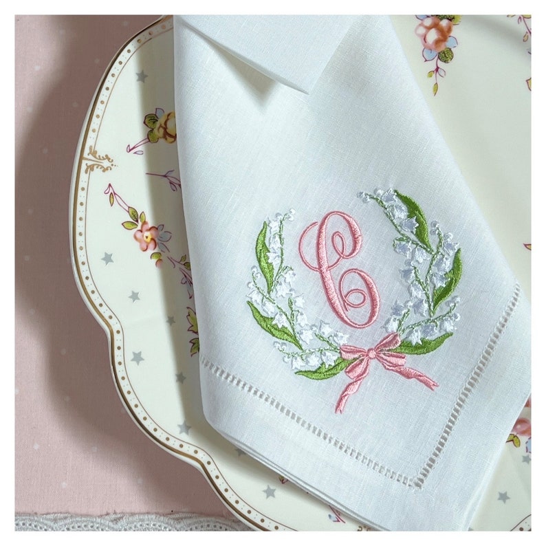 May include: White linen napkin with an embroidered wreath of lily of the valley and a pink letter "C". The wreath is green and white, with a pink bow at the bottom. The napkin has a decorative stitched border.