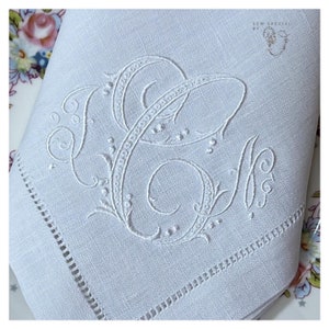 May include: White linen napkin with embroidered monogram. The monogram features the letters "C" and "M" in an elegant, ornate script. The napkin has a decorative stitched border and is folded on a floral patterned plate.
