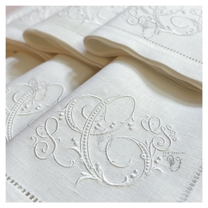 May include: A stack of white linen napkins with embroidered initials and decorative flourishes. The napkins feature a delicate, detailed design with a scalloped edge and are neatly folded. The embroidery is in a matching white thread.