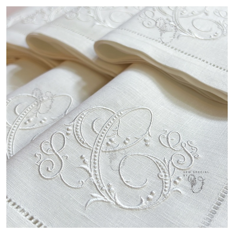 May include: A stack of white linen napkins with embroidered initials and decorative flourishes. The napkins feature a delicate, detailed design with a scalloped edge and are neatly folded. The embroidery is in a matching white thread.