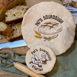May include: A beige sourdough bread cover and starter lid with embroidered text and illustrations. The larger cover reads "Pat's Sourdough" with a bread loaf and wheat design. The smaller lid says "Pat's Sourdough Starter" with a duck illustration.