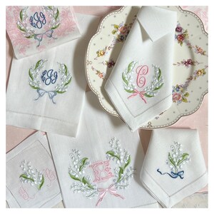 May include: White linen napkins and towels with embroidered floral designs. The embroidery includes green leaves, white flowers, and bows in pink and blue. Some pieces feature monogrammed initials.