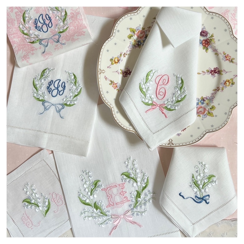 May include: White linen napkins and towels with embroidered floral designs. The embroidery includes green leaves, white flowers, and bows in pink and blue. Some pieces feature monogrammed initials.