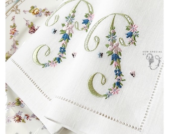 May include: Two white linen napkins with embroidered floral letters "M" in green, blue, and pink. The napkins have a scalloped edge and are folded in half. The text "Sew Special by" is embroidered in the bottom right corner.