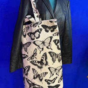 Könnte beinhalten: Eine cremefarbene Canvas-Tragetasche mit einem sich wiederholenden schwarzen Schmetterlingsdruck. Die Tasche hat zwei Schulterriemen und eine rechteckige Form. Die Schmetterlingsillustrationen variieren in Größe und Muster auf der gesamten Vorderseite.