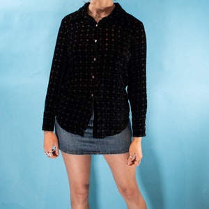 May include: A woman wearing a black velvet button-down shirt with a small geometric pattern and a denim mini skirt. She is wearing black sneakers and has curly brown hair.