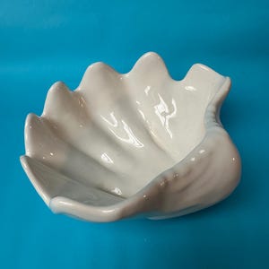 Może przedstawiać: Biała, ceramiczna miska w kształcie muszli z błyszczącym wykończeniem. Misa ma falisty, karbowany brzeg i gładkie wnętrze. Misa jest umieszczona na jasnoniebieskim tle. Ta dekoracyjna miska jest idealna do wystroju nadmorskiego.