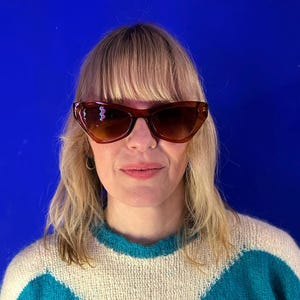 May include: Brown cat-eye sunglasses with a gradient tint. The sunglasses are worn by a person with blonde hair and a blue, white, and teal jumper. The background is a solid blue.