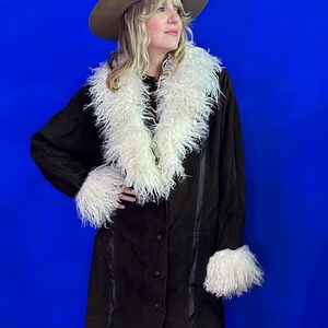 May include: A brown suede coat with a white faux fur collar and cuffs. The coat has a button closure and a long, fitted silhouette.