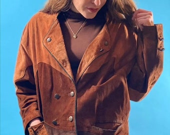 May include: A brown suede jacket with a double-breasted closure and a contrasting brown suede panel on the bottom. The jacket has two flap pockets and a zipper closure.