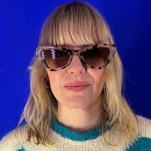May include: Cat-eye sunglasses in a tortoiseshell pattern with brown gradient lenses. The sunglasses feature a wavy neon pink and blue design on the left lens. The person is wearing a teal and white sweater.
