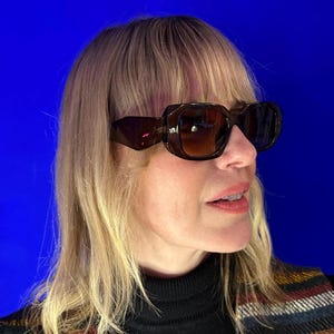 Puede incluir: Gafas de sol marrones de gran tamaño con montura cuadrada. Las gafas de sol tienen un tinte oscuro y las lleva una persona con cabello rubio. El fondo es azul liso.