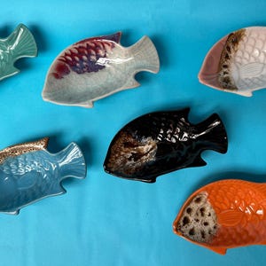 Può includere: Una collezione di sei piatti in ceramica a forma di pesce in vari colori, tra cui verde, bianco, blu, nero e arancione. Ogni piatto presenta un motivo a scaglie strutturato e un distinto dettaglio di smalto marrone maculato vicino alla testa.
