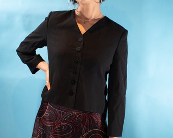 Puede incluir: Una mujer con un blazer negro con botones y una falda de estampado negro y rojo. Tiene el pelo castaño rizado y lleva pendientes de aro plateados.