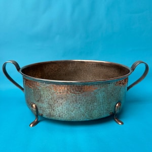 May include: A round, hammered metal bowl with a tarnished silver and copper-toned finish. The bowl features two curved handles on opposite sides and is supported by three small, delicate feet extending from the base of the vessel.