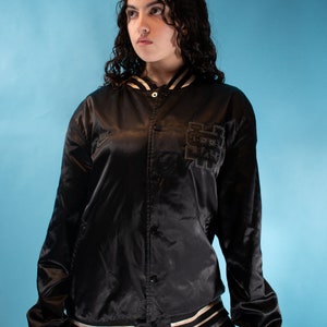 May include: A black satin bomber jacket with a white and black striped trim. The jacket has a button closure and a large embroidered patch on the left side. The patch features a stylized graphic with the text "NYC".