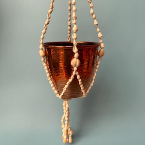 Könnte beinhalten: Ein gehämmertes, kupferfarbenes Metallpflanzgefäß, das in einem Makramee-artigen Aufhänger aus kleinen, cremeweißen Muscheln hängt. Der Aufhänger besteht aus mehreren Muschelsträngen, die den schalenförmigen Behälter umschließen.