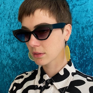 May include: Black cat-eye sunglasses with gradient lenses. The person is wearing yellow lemon slice earrings and a black and white patterned shirt. The background is a textured blue.