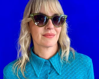 Könnte beinhalten: Eine Frau mit blonden Haaren, die eine grüne Cat-Eye-Sonnenbrille und ein blaues Hemd mit Kragen und einem strukturierten Muster trägt.