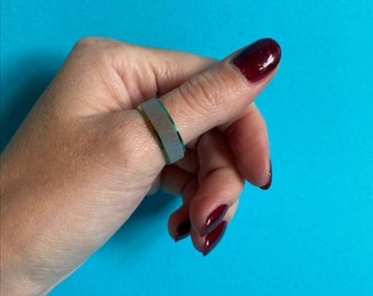 May include: A silver ring with a green and gold band on a finger with dark red nail polish.