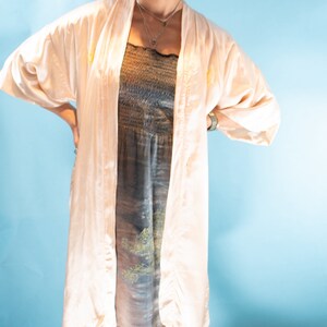 May include: A woman wearing a white satin robe over a patterned dress. She has curly brown hair and is wearing black sneakers.