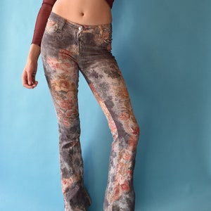 Può includere: Un paio di jeans a zampa di elefante grigi con una stampa floreale con fiori rosa, rossi e bianchi. I jeans sono indossati da una persona con una maglia rossa su uno sfondo blu.