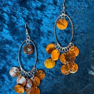 May include: A pair of dangle earrings with silver-tone metal oval frames. Each earring features a single, round, brown and gold-toned disc at the top, with multiple smaller, round, brown and gold-toned discs dangling below. The earrings are set against a blue background.