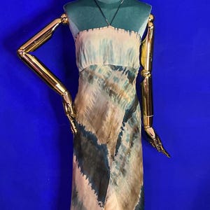 May include: A strapless, floor-length dress with a tie-dye pattern in shades of blue, beige, and brown. The dress is displayed on a mannequin with a teal head and gold arms. A simple necklace is visible.