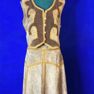 Può includere: Un completo vintage a due pezzi composto da gilet e gonna. Il gilet marrone presenta dettagli beige e bottoni. La gonna beige ha finiture marroni e dorate. L'outfit è esposto su un manichino su sfondo blu.