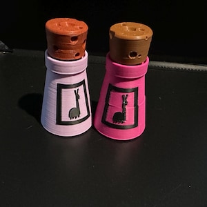 Pode incluir: Dois recipientes cónicos com tampas castanhas semelhantes a cortiça. Um recipiente é cor-de-rosa claro e o outro é cor-de-rosa escuro. Cada um apresenta um rótulo retangular preto na frente com a silhueta preta de uma lhama virada à esquerda.