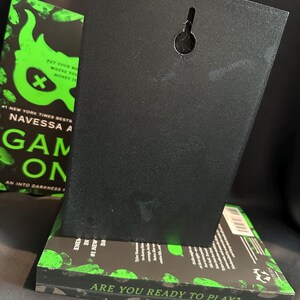 K&ouml;nnte beinhalten: Eine schwarze, rechteckige Wandtafel mit einem Schl&uuml;sselloch zum Aufh&auml;ngen, die auf einem Buch ruht. Das Buch hat einen schwarz-gr&uuml;nen Einband mit dem Text "GAME ON" und "ARE YOU READY TO PLAY?" Der Hintergrund ist dunkel.