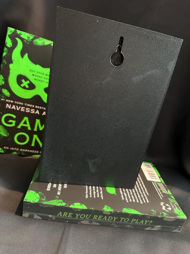 K&ouml;nnte beinhalten: Eine schwarze, rechteckige Wandtafel mit einem Schl&uuml;sselloch zum Aufh&auml;ngen, die auf einem Buch ruht. Das Buch hat einen schwarz-gr&uuml;nen Einband mit dem Text "GAME ON" und "ARE YOU READY TO PLAY?" Der Hintergrund ist dunkel.